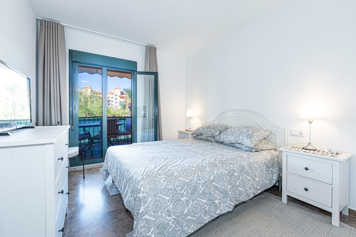 A bright bedroom with a double bed, bedside tables, a dresser, and a balcony with outdoor seating and a view of buildings and trees.