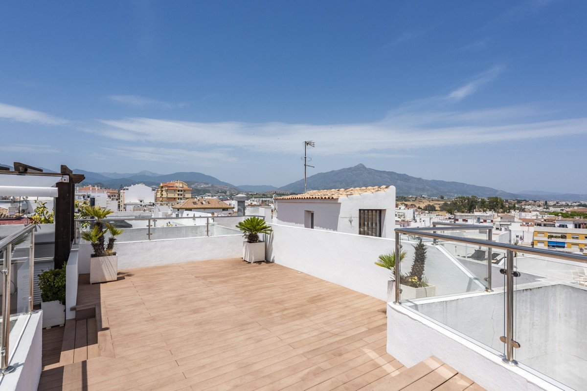 Rooftop terrace with wooden deck, glass railings, potted plants, and mountain views under a clear blue sky.