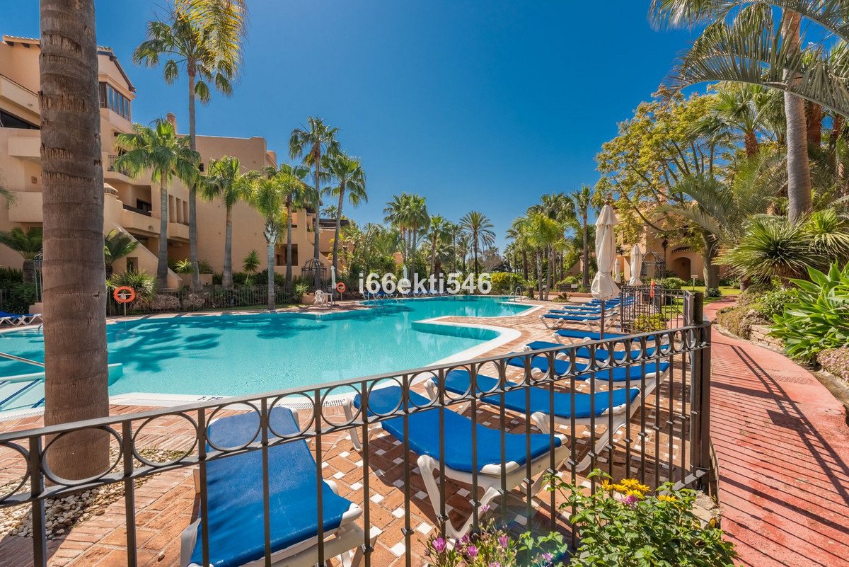 A resort swimming pool with palm trees, sunbeds, and umbrellas, under a clear blue sky.