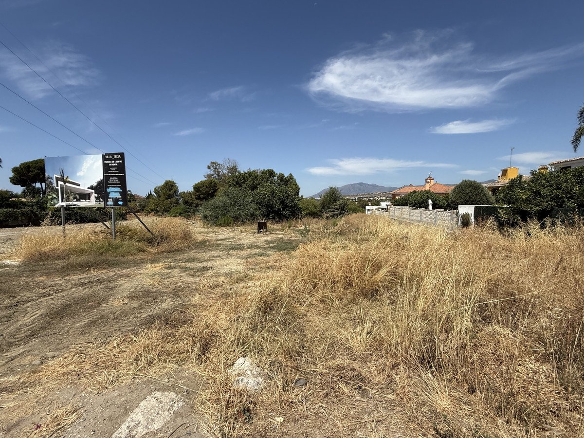 A sign for a villa stands in a vacant plot of land with dry grass and distant buildings under a blue sky.