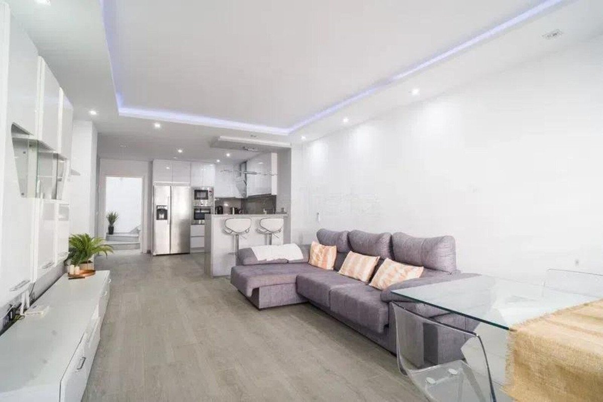 Modern open-plan living area with a grey sectional sofa, breakfast bar, kitchen appliances, and a glass dining table.