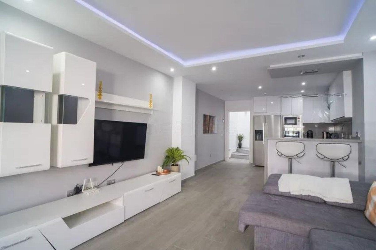 Modern living area with wall-mounted TV, sleek cabinets, and a view of a kitchenette with bar stools.