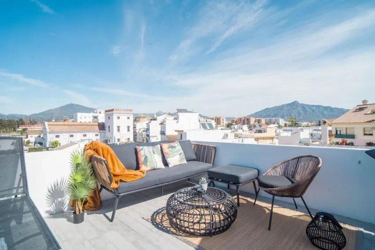 A modern terrace with a sofa, chairs, and coffee table offering a view of town and mountains under a clear blue sky.