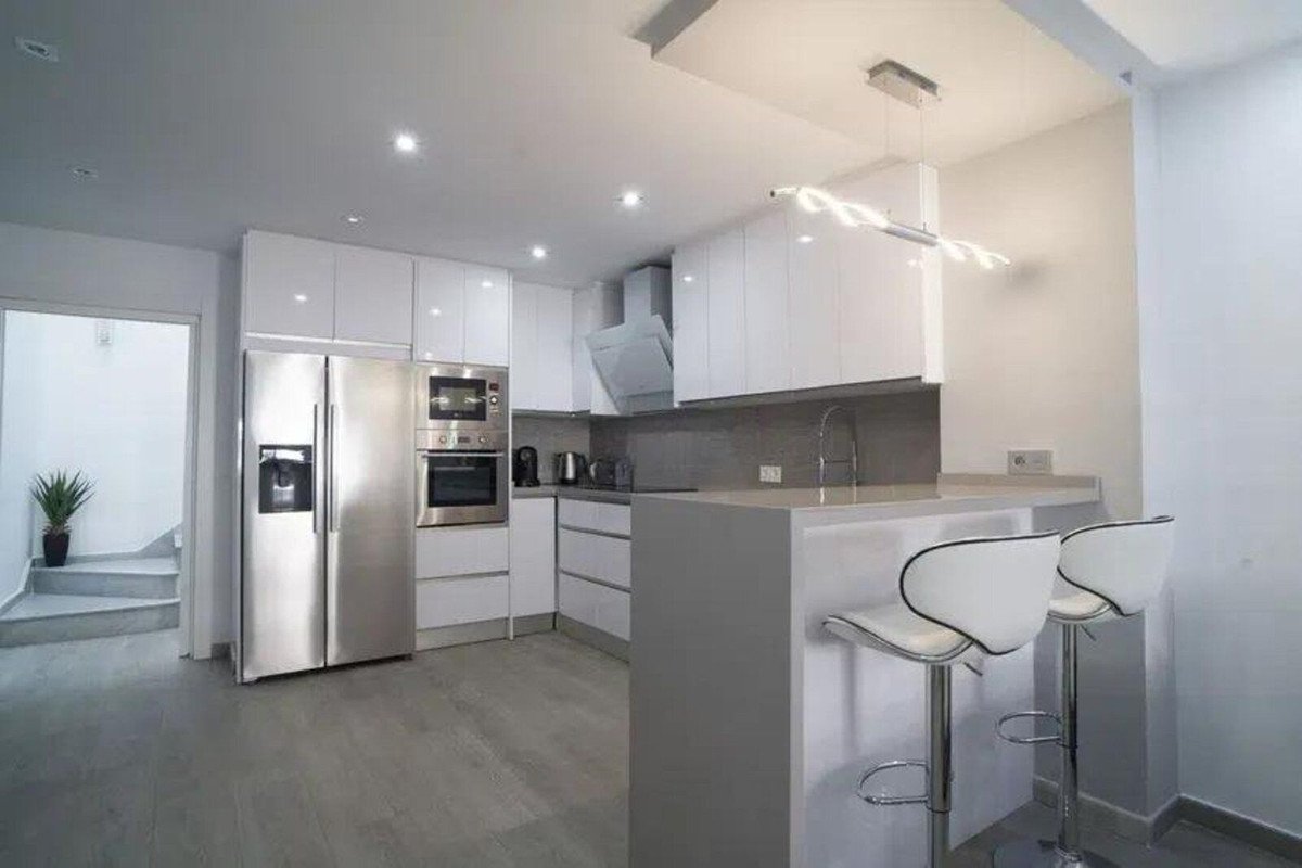 Modern white kitchen with stainless steel appliances, breakfast bar, and two stylish stools. View shows stairs to the left.
