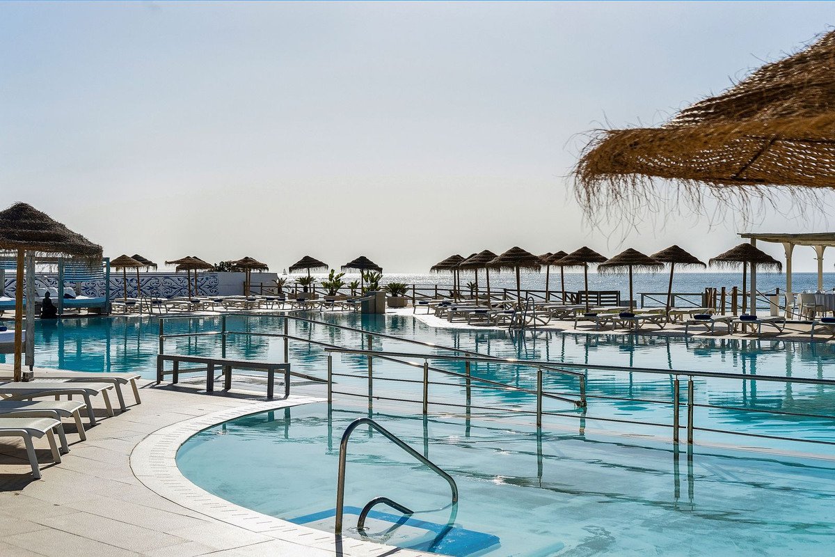 Large outdoor swimming pool area with lounge chairs, umbrellas, and a view of the ocean.