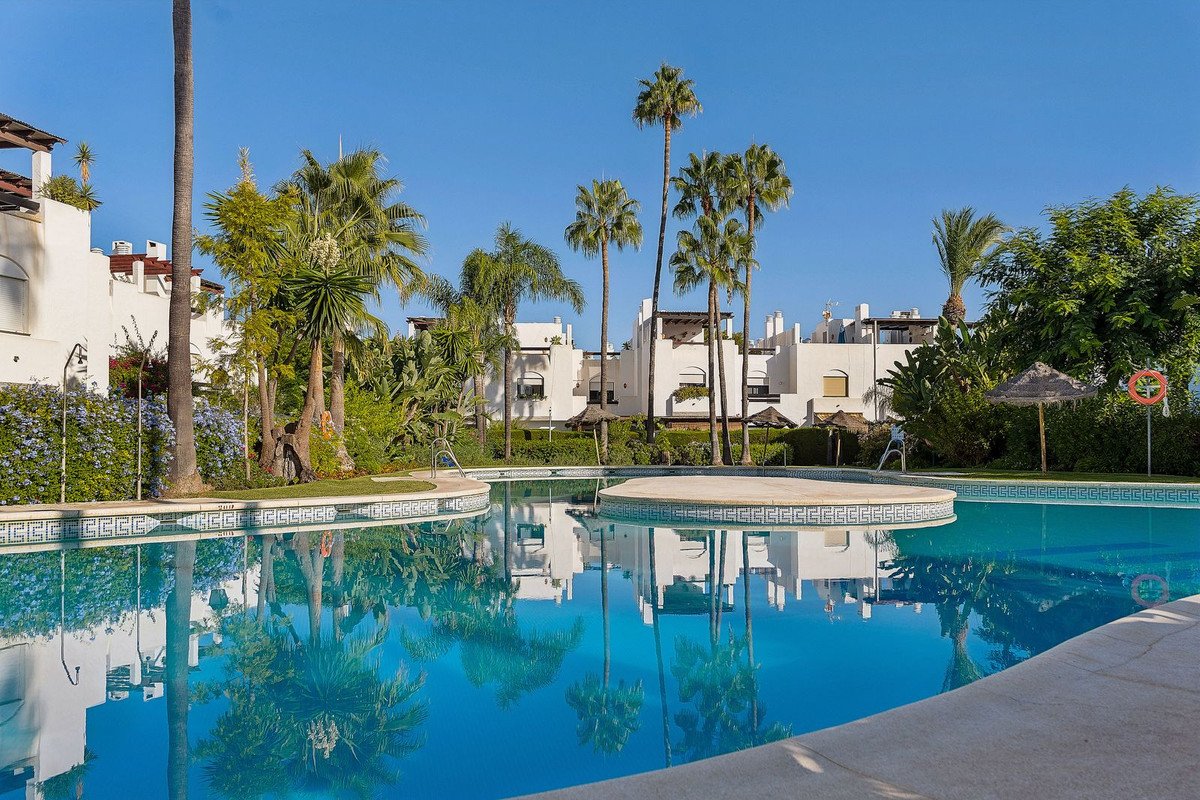 A sparkling blue swimming pool surrounded by lush green palm trees and white buildings under a clear blue sky.