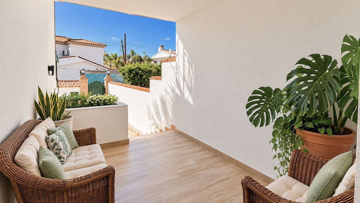 A sunny terrace with wicker seating, potted plants, and a view of neighboring houses and greenery.