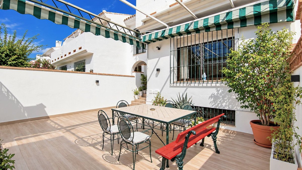 Outdoor terrace with a table, chairs, and bench, featuring potted plants and awnings on a sunny day.