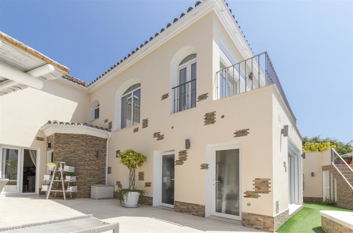 Exterior view of a modern villa with a balcony, patio, and sunny blue sky. Features include arched windows and stone accents.