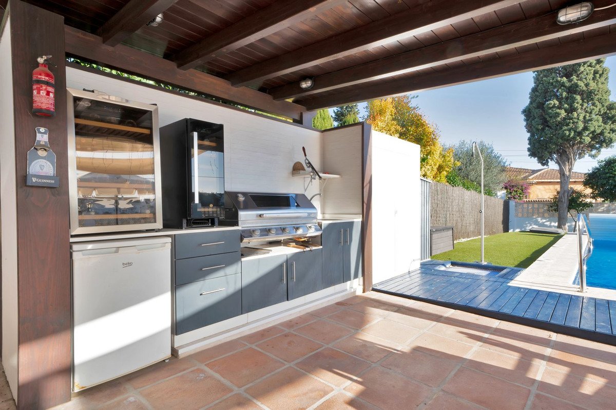 Covered outdoor kitchen area with a grill, wine cooler, and refrigerator, next to a swimming pool and artificial turf.