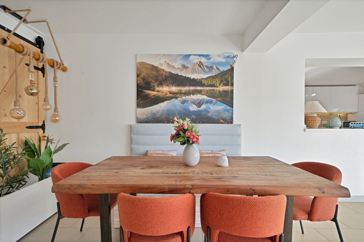 A dining area with a wooden table, orange chairs, and a large artwork of a mountain lake scene above a headboard.