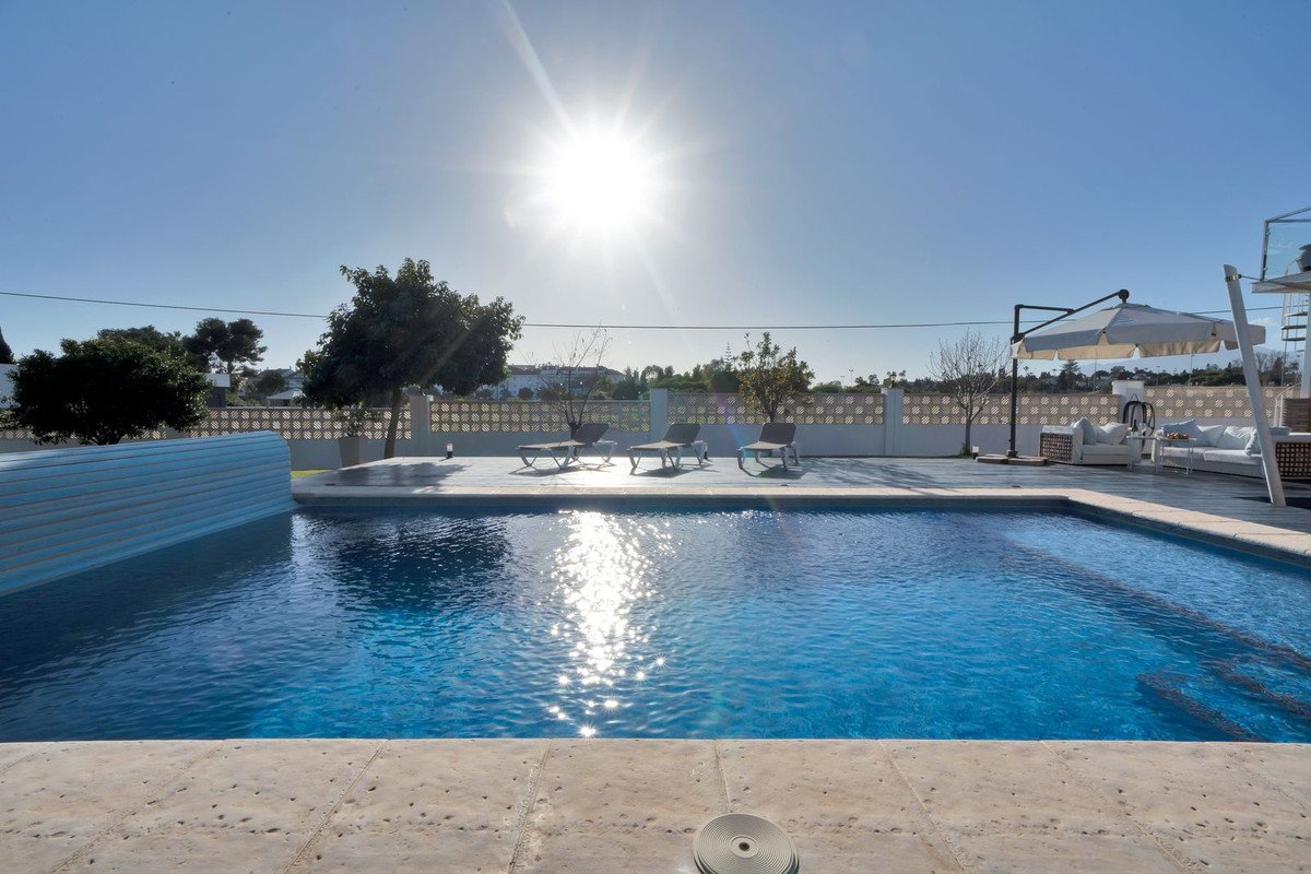 Sunny day by a large blue swimming pool, with lounge chairs, sofas, and umbrellas on the patio.