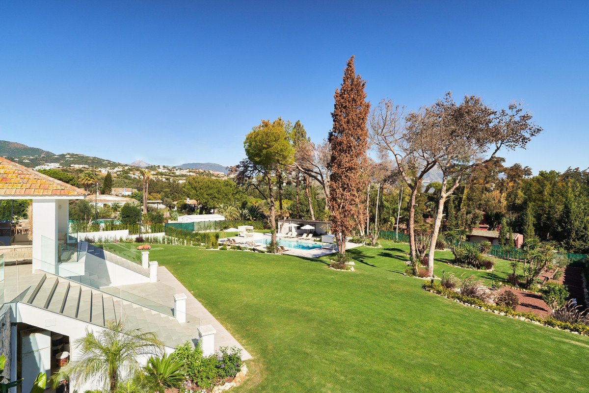 Spacious villa with a swimming pool and manicured lawn, set against a backdrop of green hills and mountains under a clear blue sky.