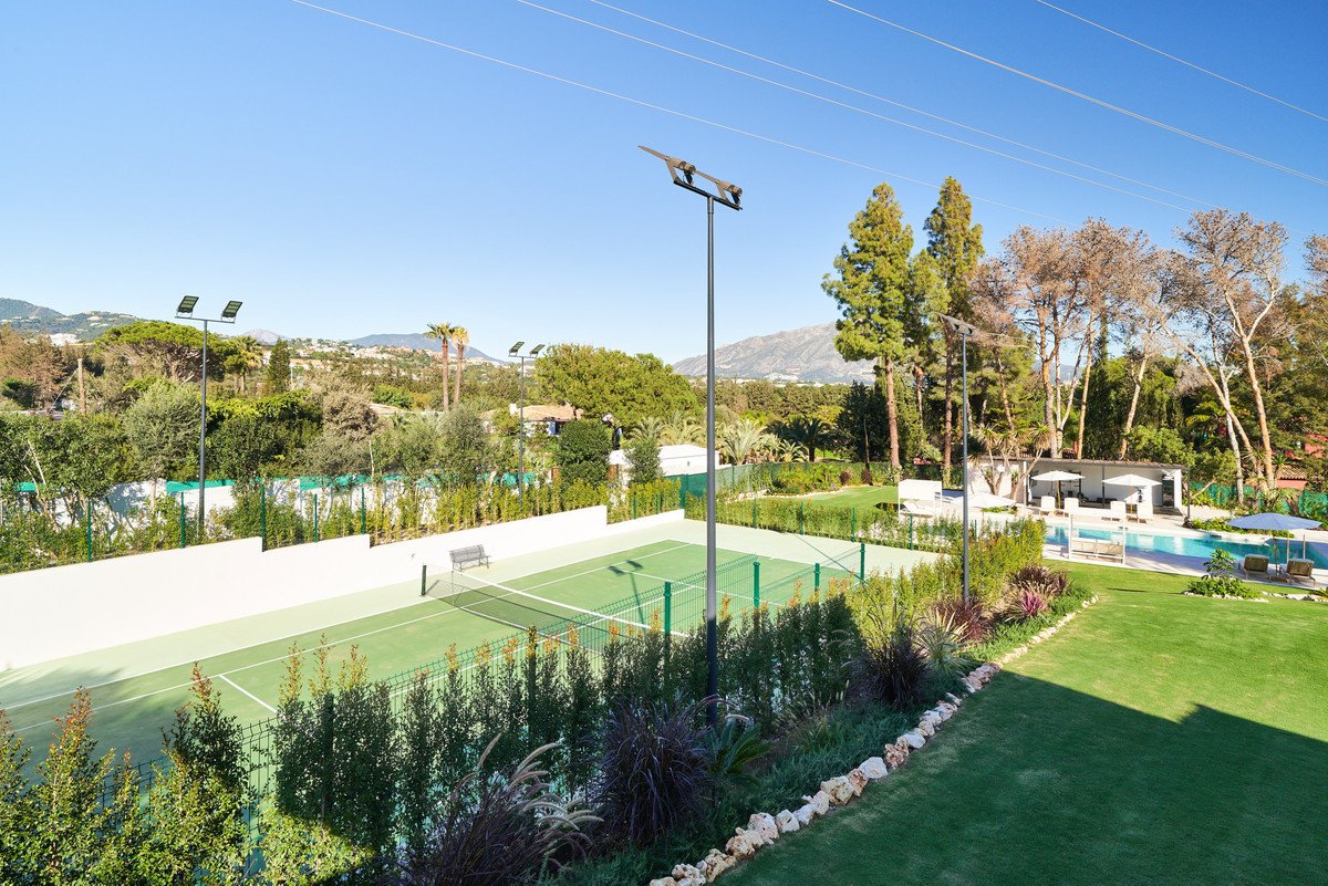 A tennis court and swimming pool area on a sunny day, surrounded by lush greenery and trees with a mountain view.