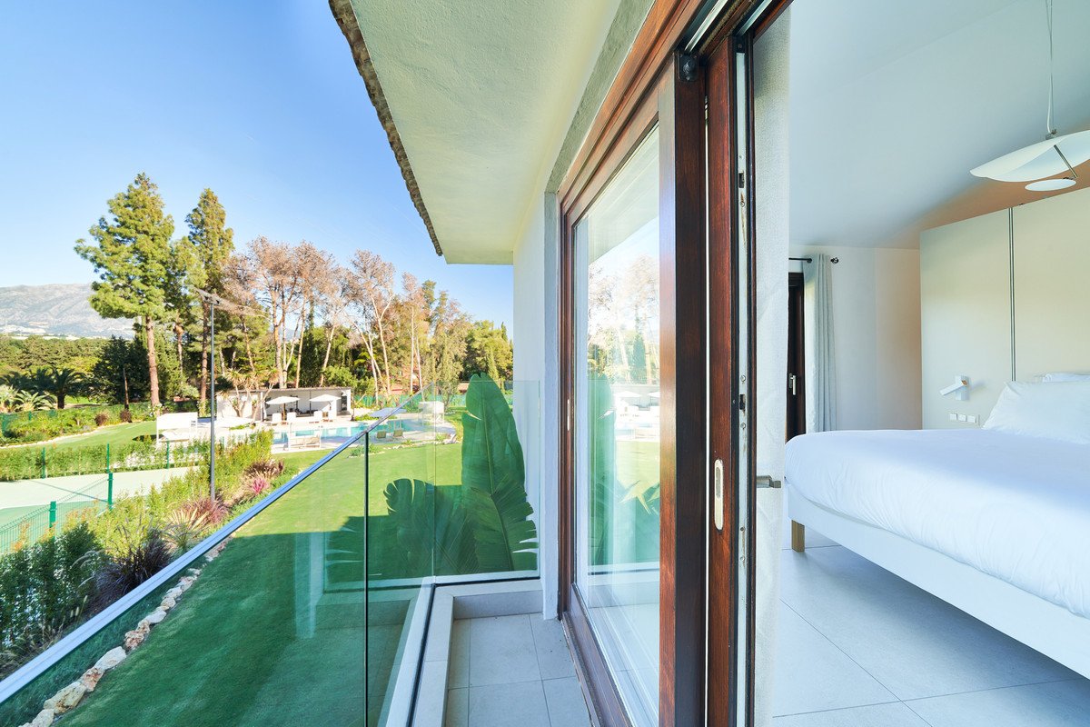 View from a balcony with a swimming pool and golf course beyond, overlooking lush greenery and trees under a clear blue sky.
