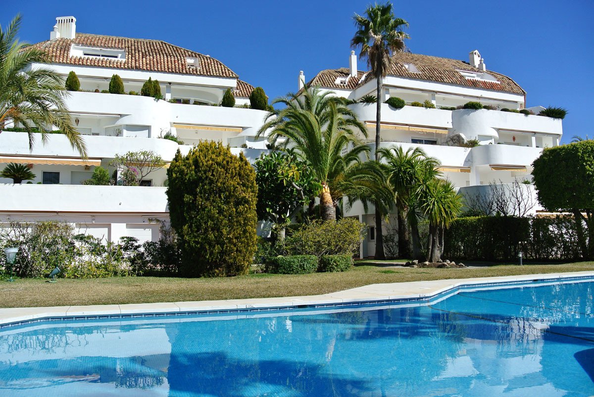 A white apartment building with balconies and tiled roofs surrounded by palm trees, overlooking a bright blue swimming pool.