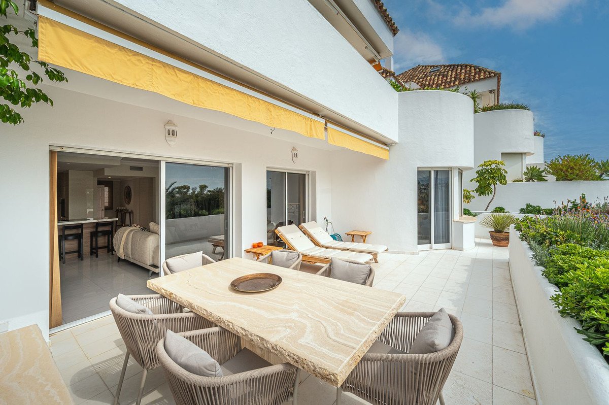 Spacious modern terrace with dining table, lounge chairs, and greenery, overlooking a living room and distant trees.