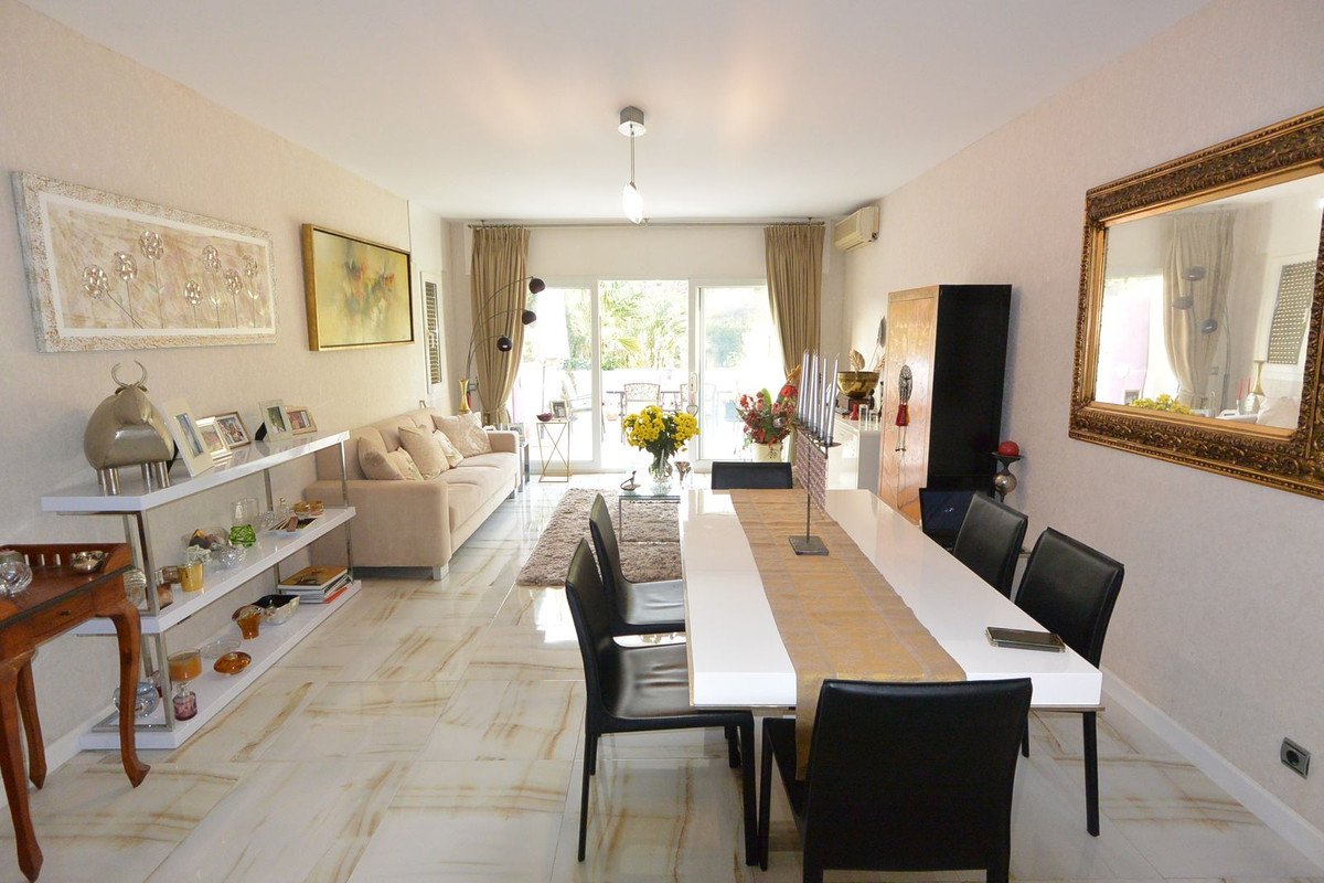 A spacious living and dining room with a long white table, beige sofa, and a view of a sunny terrace through glass doors.