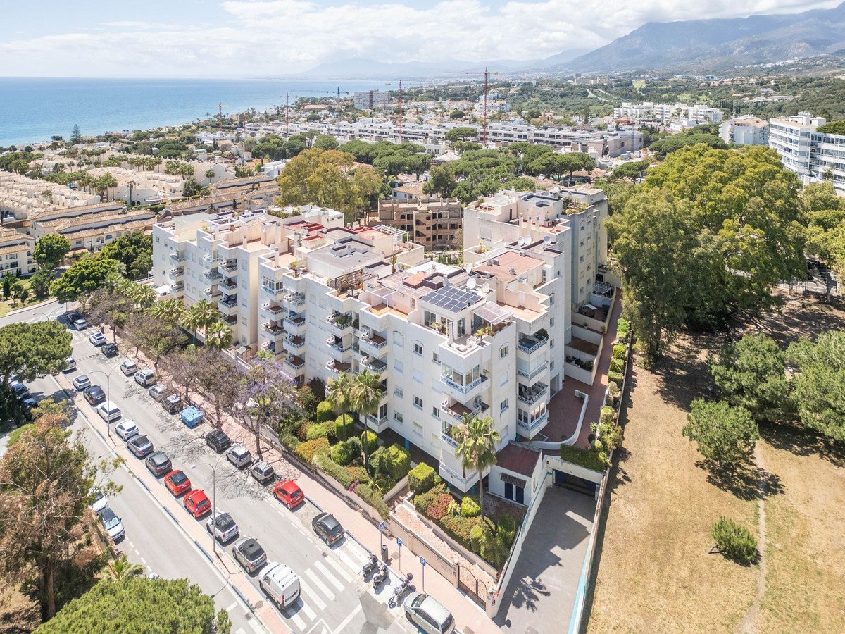 Aerial view of a white apartment building with balconies, overlooking the sea and mountains in the distance.