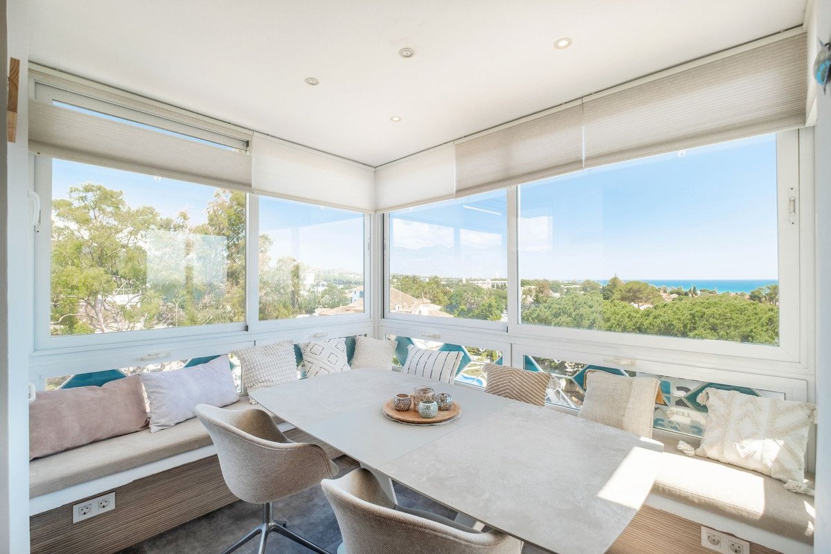 Bright dining area with built-in seating and table, overlooking lush greenery and the sea from a sunlit terrace.