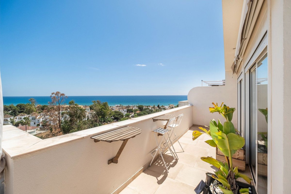 A sunny balcony with two folding chairs and tables, offering a view of the sea and coastal town below.