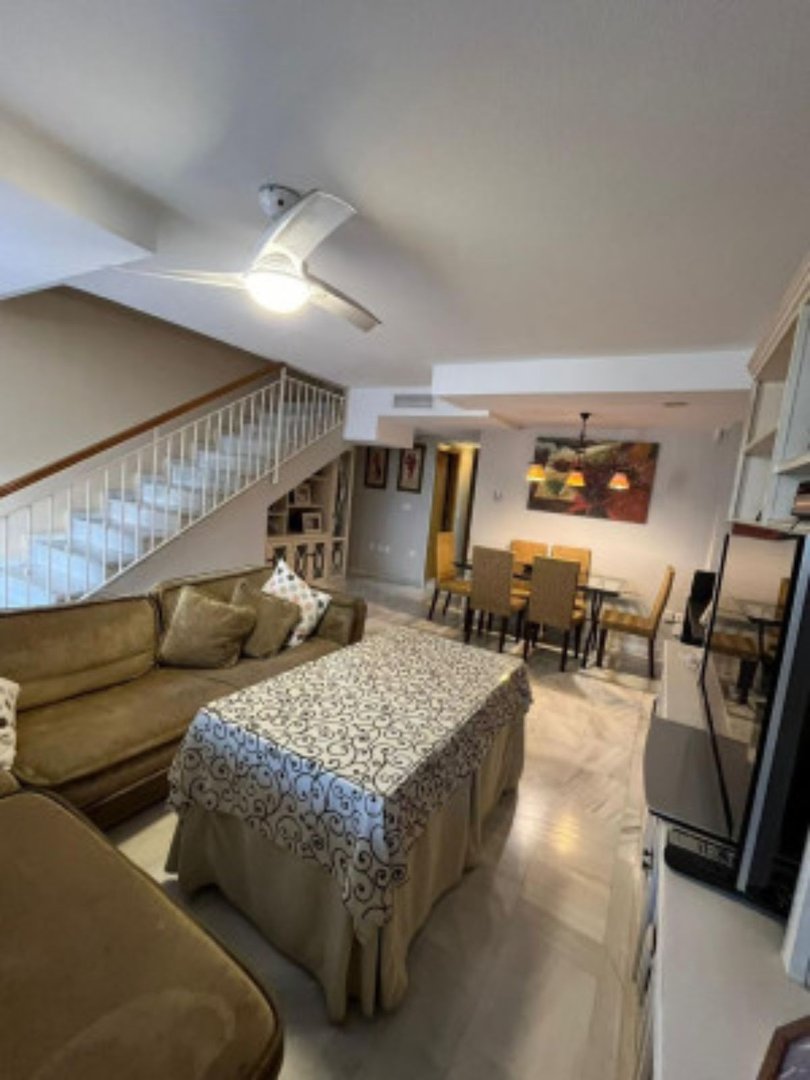 A spacious living and dining area with a large sofa, coffee table, dining set, and stairs leading upstairs.