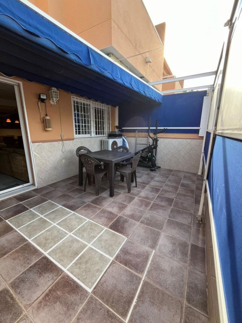 A tiled terrace with a table, chairs, and an exercise bike under a blue awning.