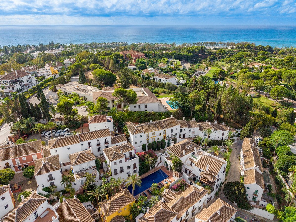 Aerial view of white villas with tiled roofs, swimming pools, lush greenery, and the ocean in the background.