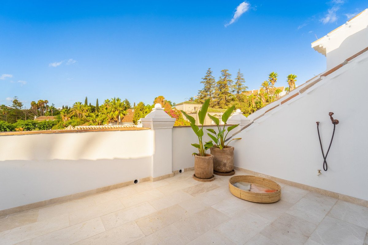 A sunny terrace with potted plants, a woven basket, and an outdoor shower, overlooking lush green hills and blue sky.