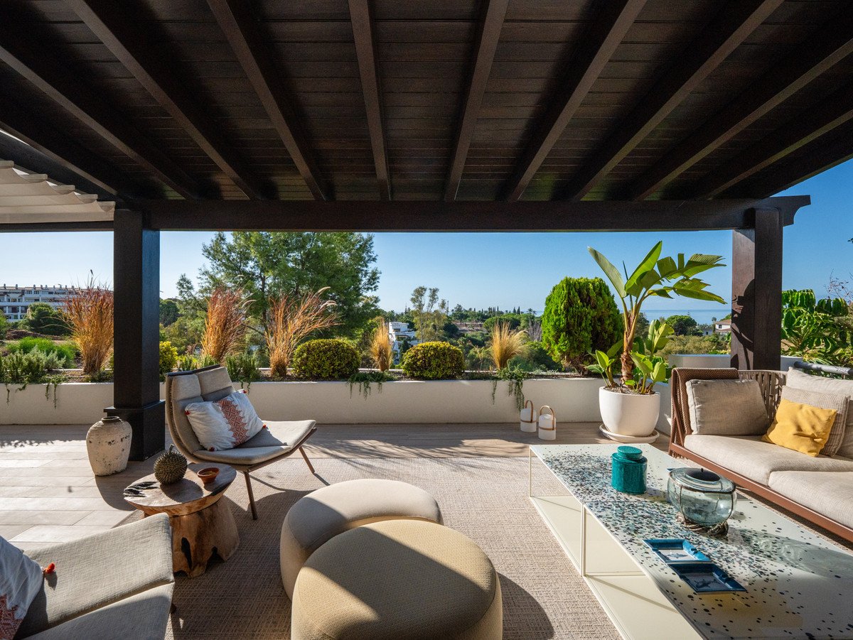A covered terrace with outdoor seating, lush greenery, and a distant sea view under a clear blue sky.