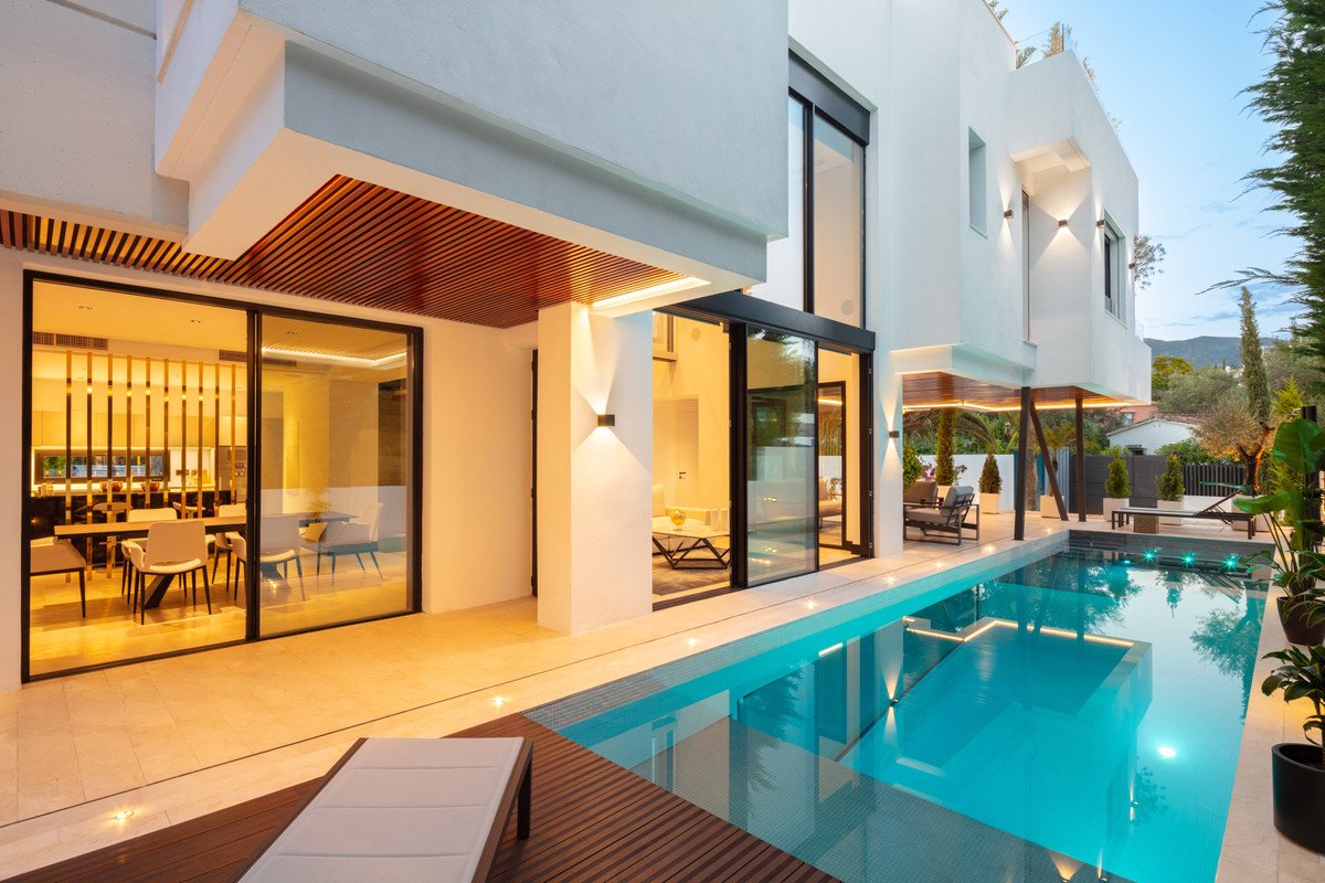 Modern villa exterior with a swimming pool, outdoor dining area, and floor-to-ceiling windows revealing an interior dining room.