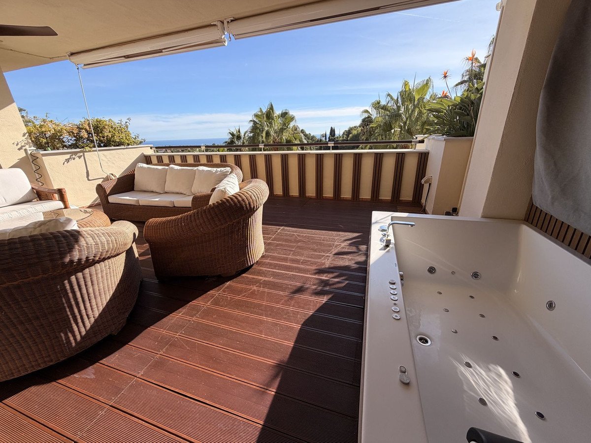 A spacious terrace with rattan seating, a jacuzzi tub, and a view of palm trees and the sea under a clear blue sky.