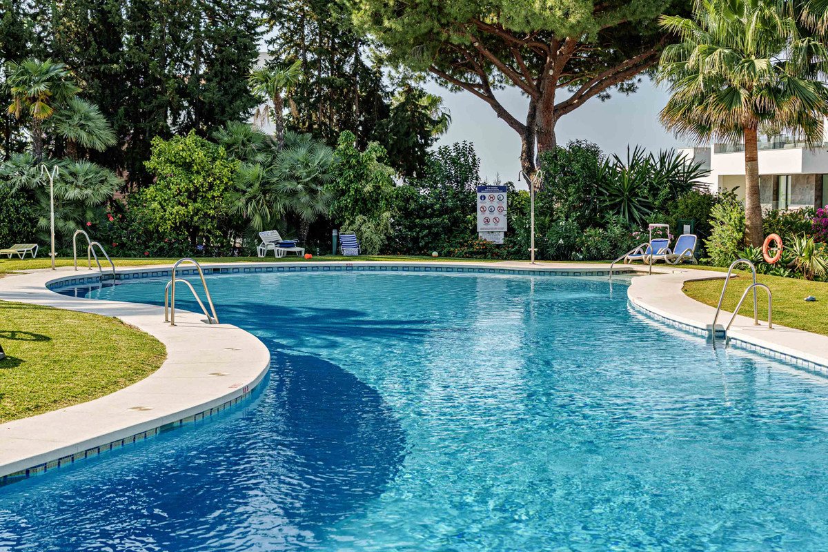 A curved swimming pool with steps and lounge chairs set in a lush, green garden with palm trees and a building in the background.