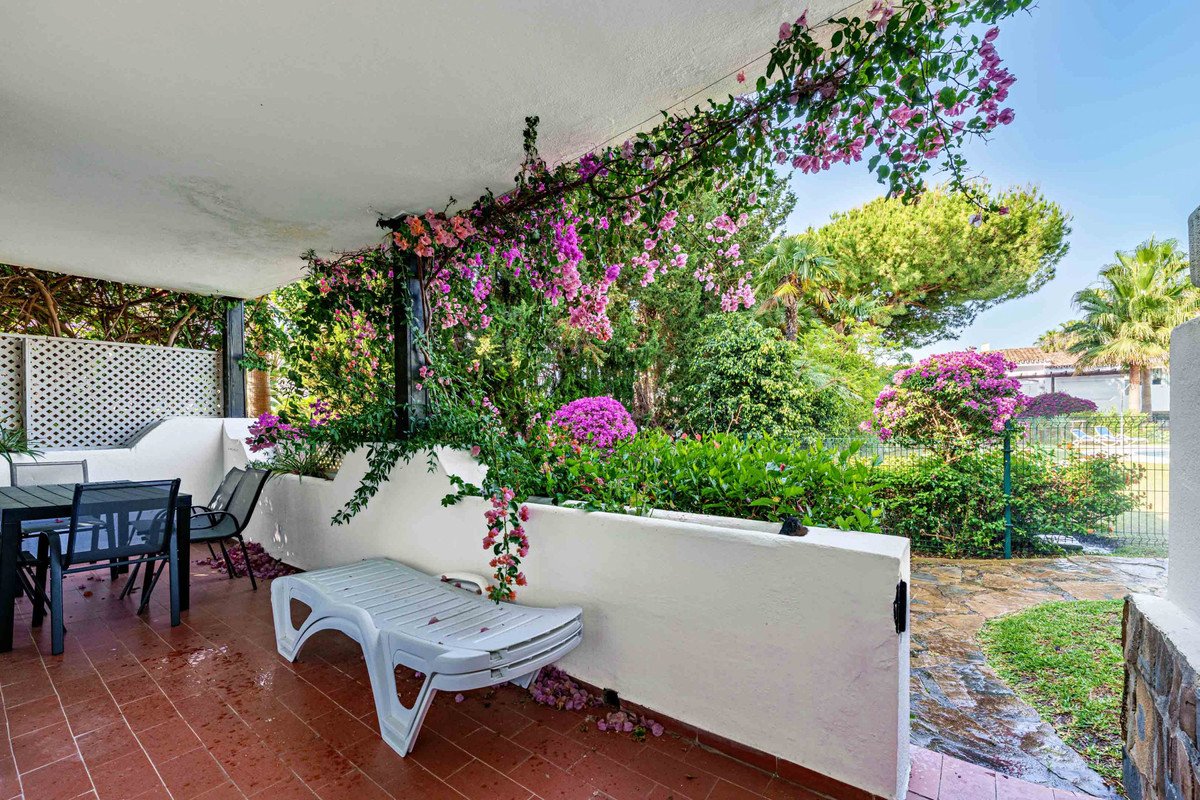 A covered terrace with a dining set, lounge chair, and abundant flowering plants overlooking a garden and trees.