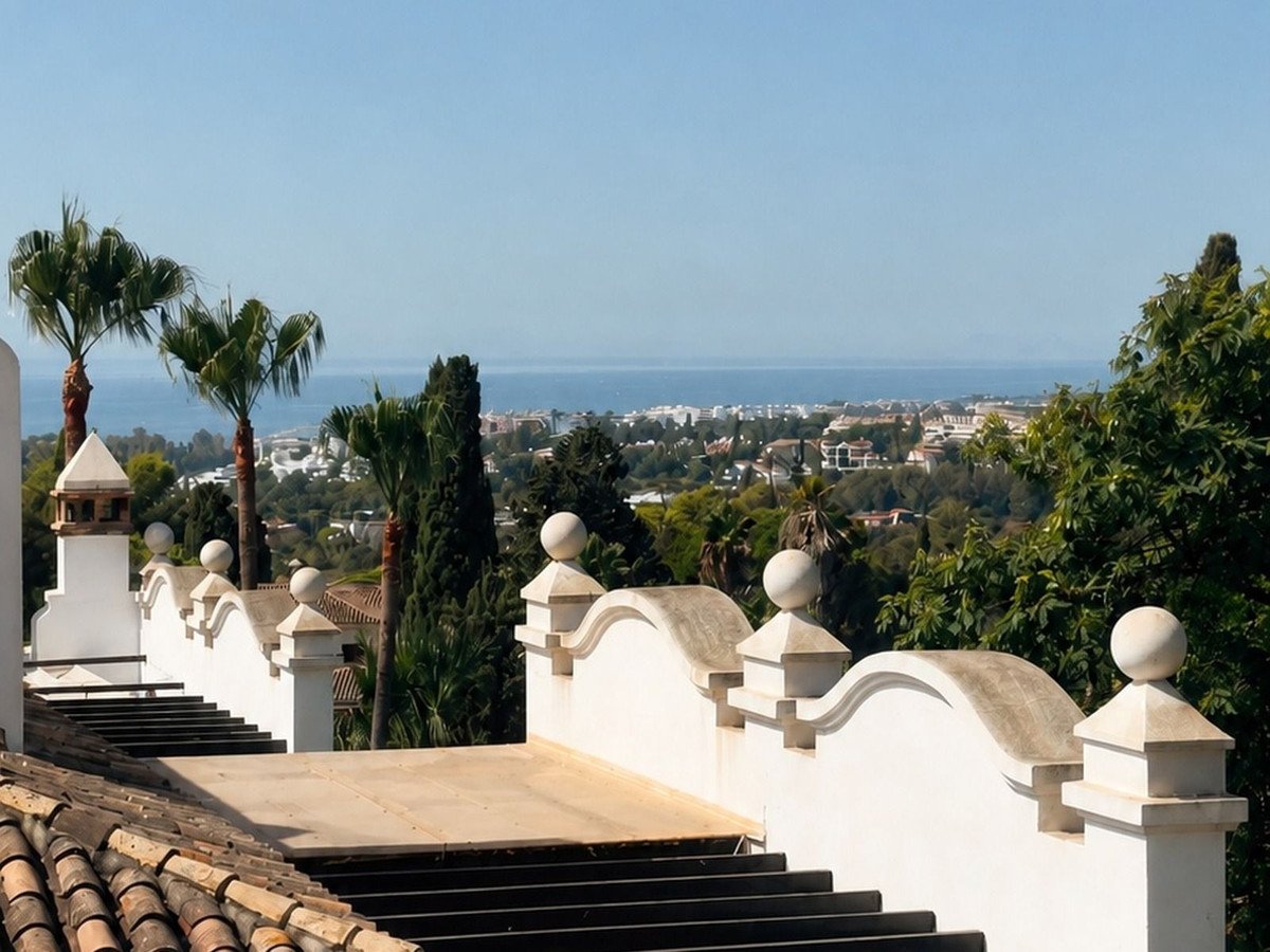 A terrace with stairs, white railings, and palm trees overlooking the sea and city.