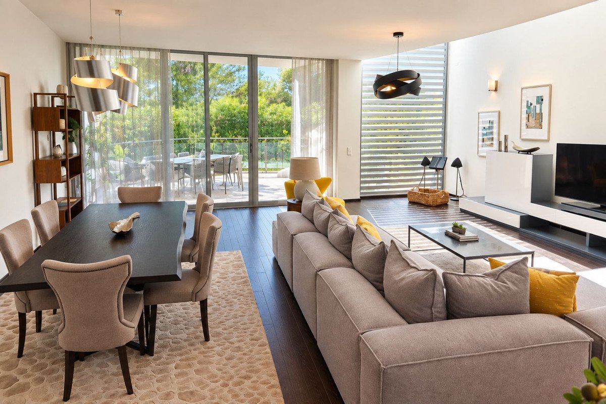 Spacious living and dining area with large windows overlooking a patio and lush green trees. Features a dining table, sofa, and modern lighting.