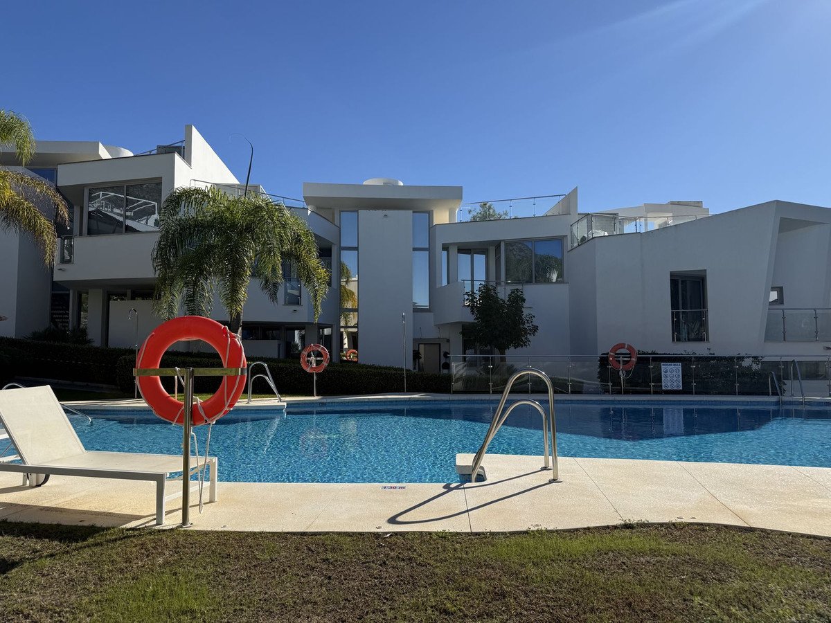 Modern white apartments with balconies overlooking a bright blue swimming pool, sun loungers, and palm trees under a clear sky.