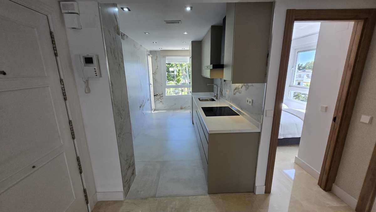 A modern kitchen with sleek countertops and an induction hob, connected to a living area with natural light. A bedroom is visible through an open doorway.