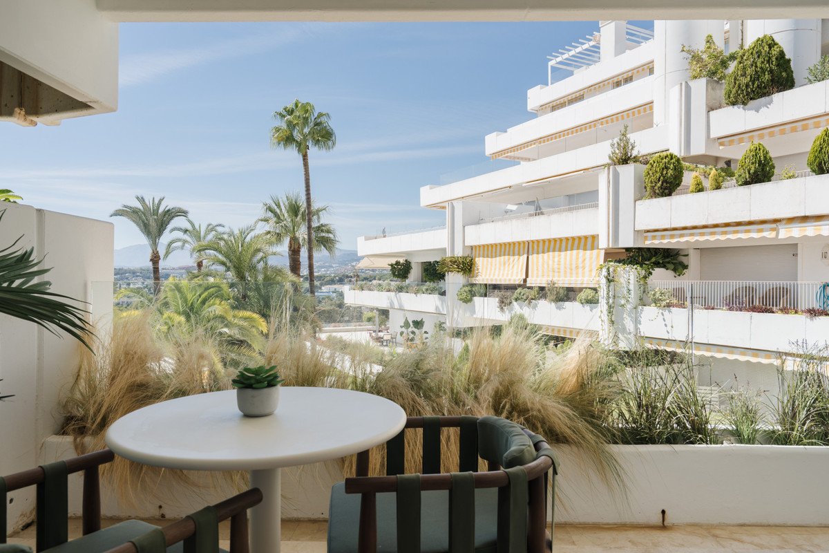 A white table and chairs on a balcony with lush greenery and a view of palm trees and a building.