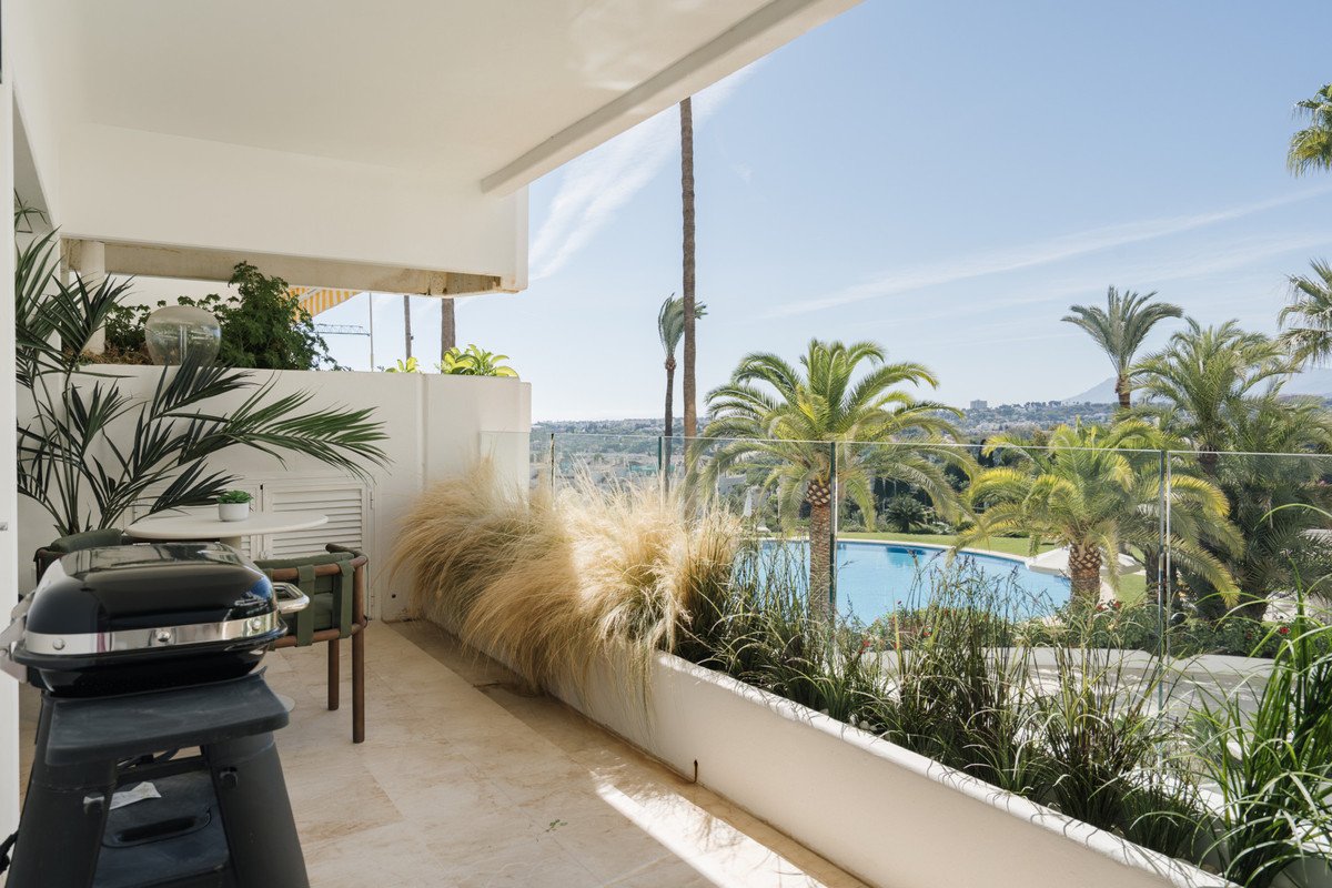 A balcony with a grill, seating area, and a view of a swimming pool and palm trees on a sunny day.