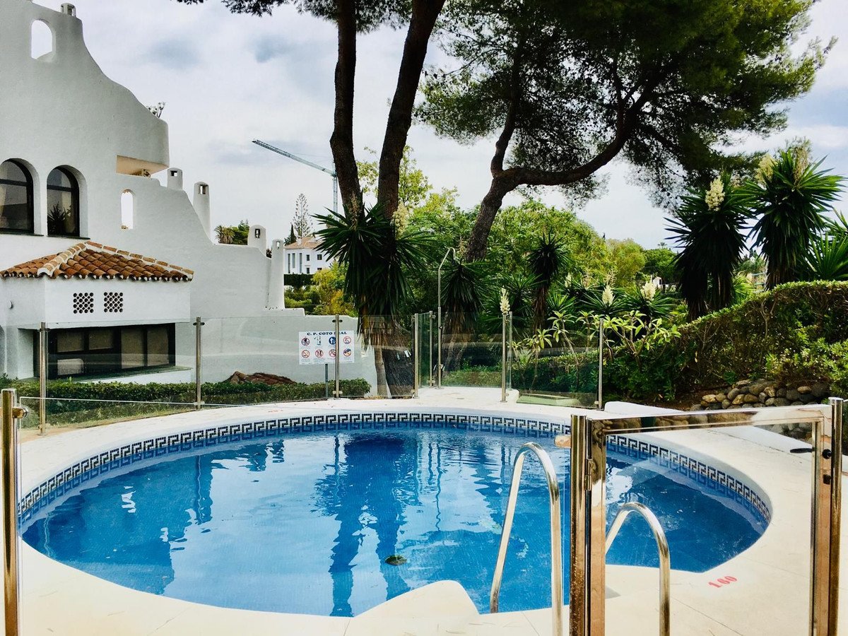 Circular swimming pool with steps and glass fence, surrounded by lush greenery and a white building.