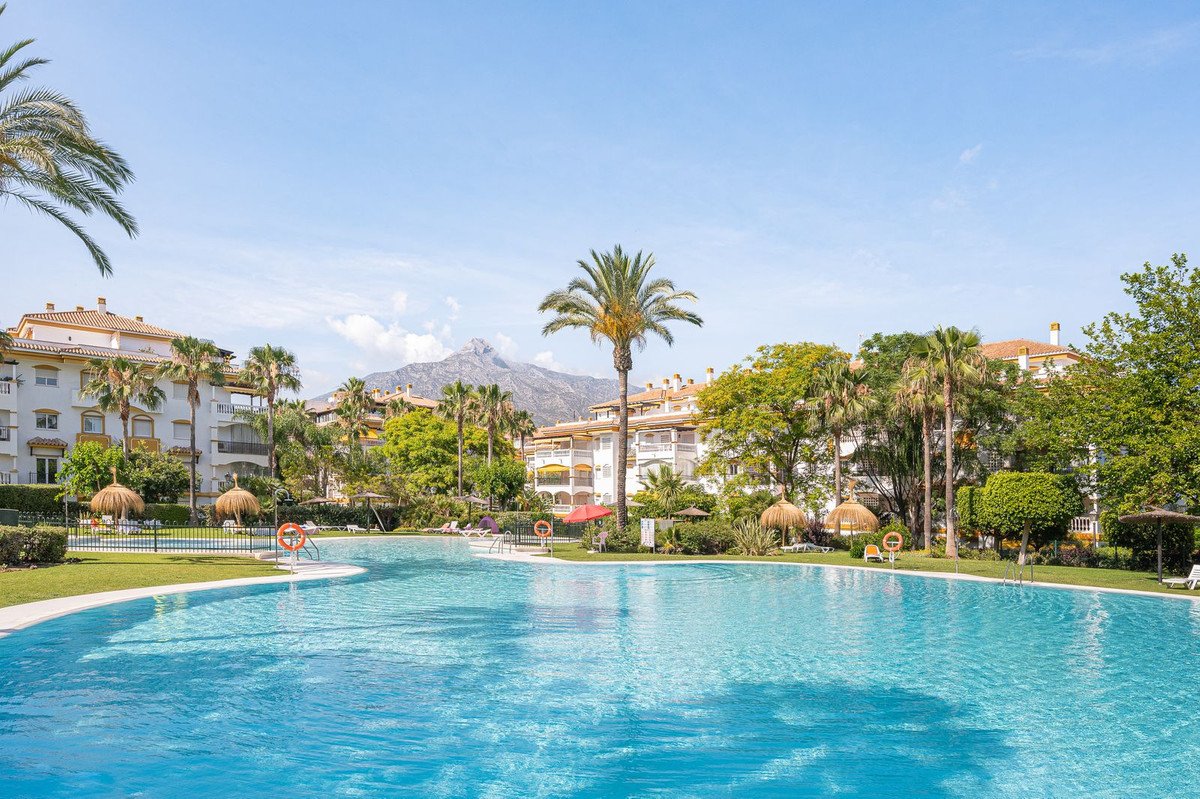 A resort complex with a large swimming pool, palm trees, and mountains in the background under a clear blue sky.