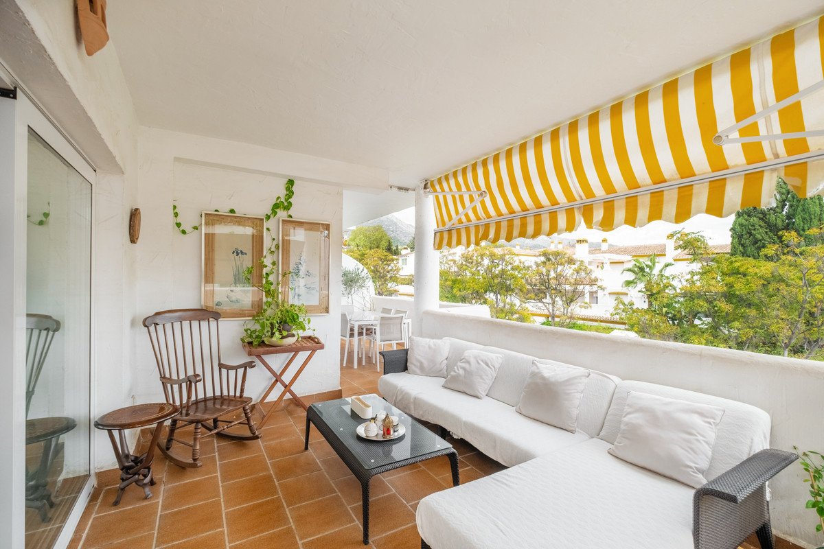 Covered terrace with white sectional sofa, coffee table, rocking chair, and a view of trees and houses under a yellow awning.