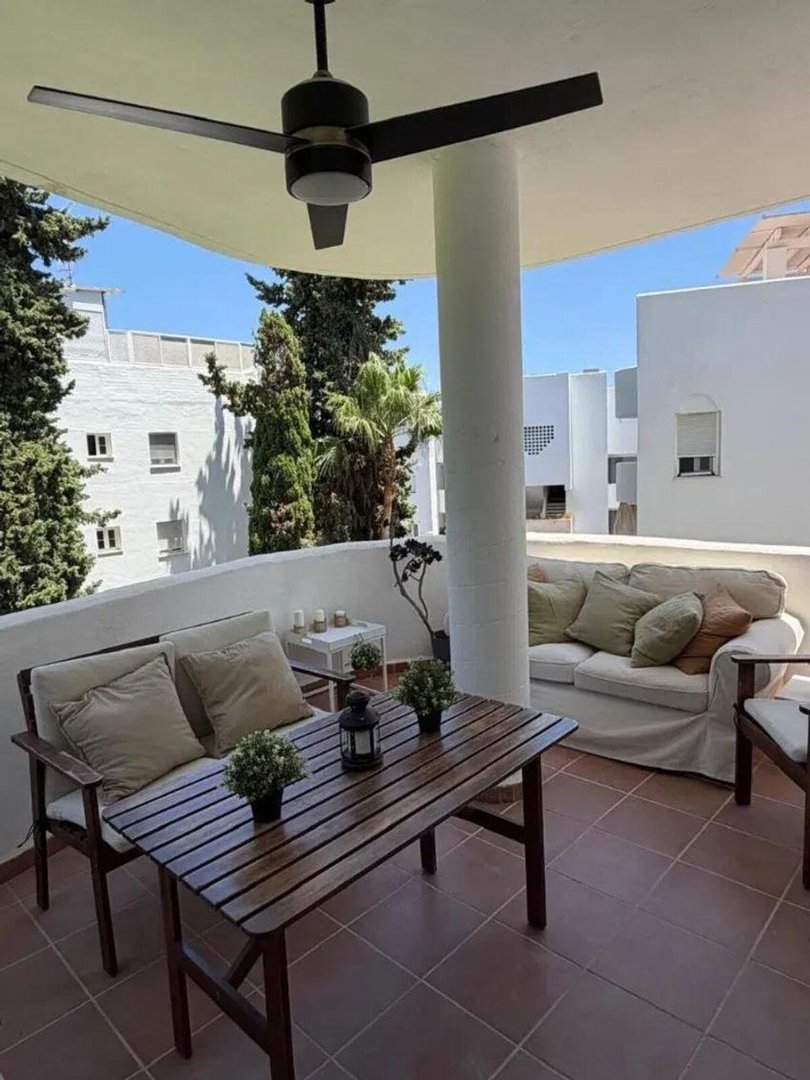 A covered balcony with outdoor seating, including a table, chairs, and a sofa, overlooking buildings and trees.