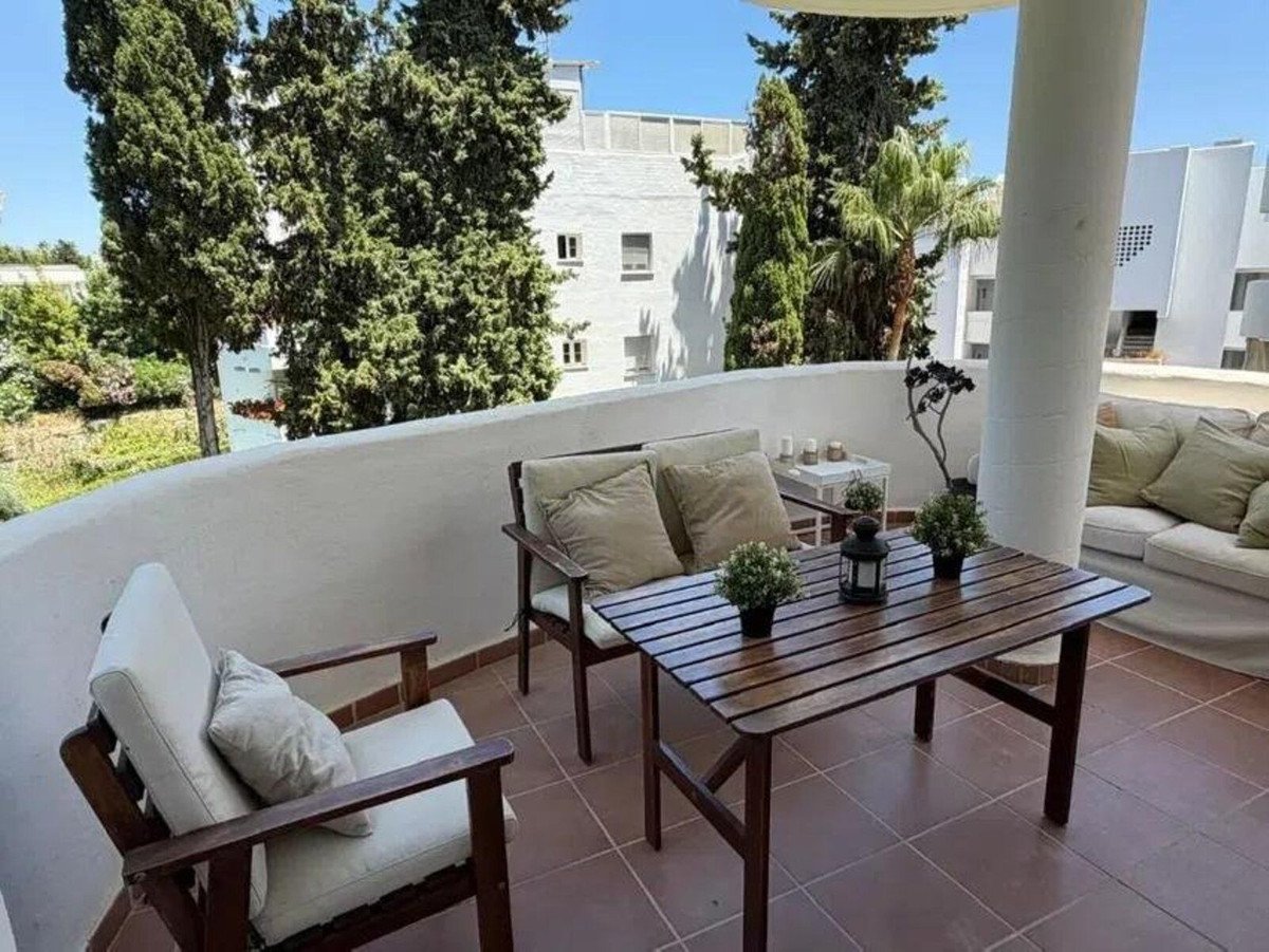 A cozy balcony seating area with a wooden table and chairs, offering a view of trees and buildings.