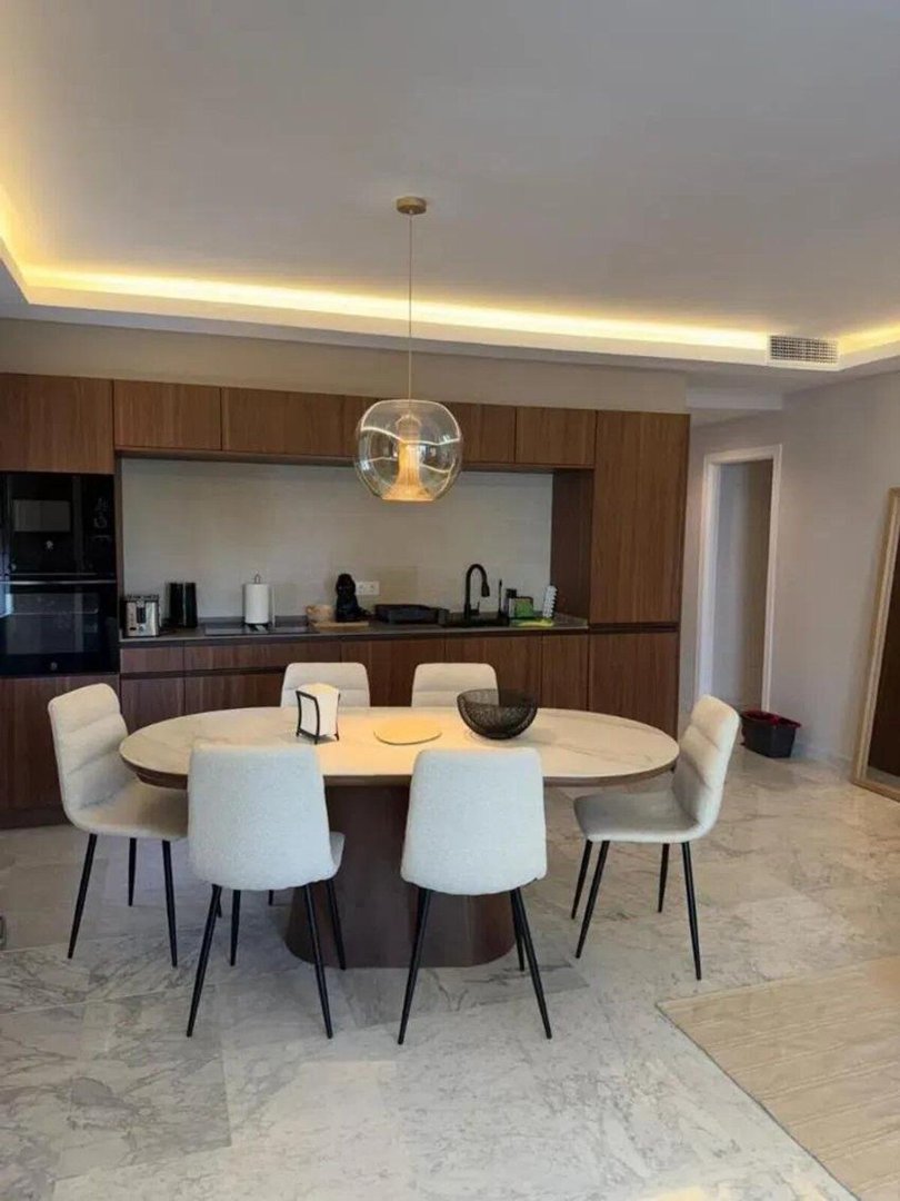 Modern dining area with an oval table and chairs, connected to a contemporary kitchen with wooden cabinets and integrated appliances.