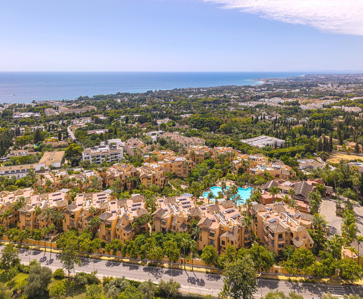 Aerial view of a residential complex with apartments, palm trees, a swimming pool, and the sea in the distance.