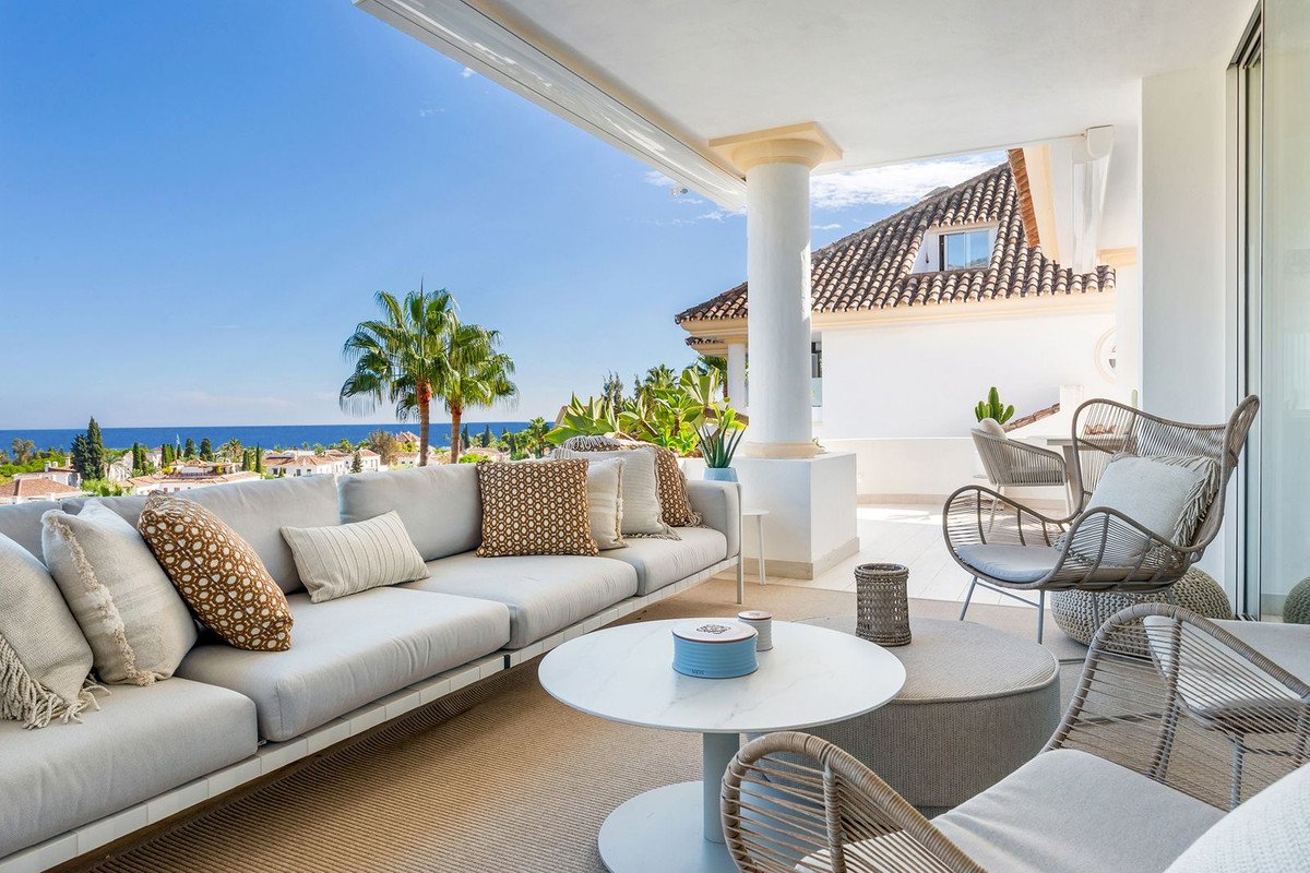 Modern terrace with comfortable seating and a beautiful sea view, featuring palm trees and white buildings.