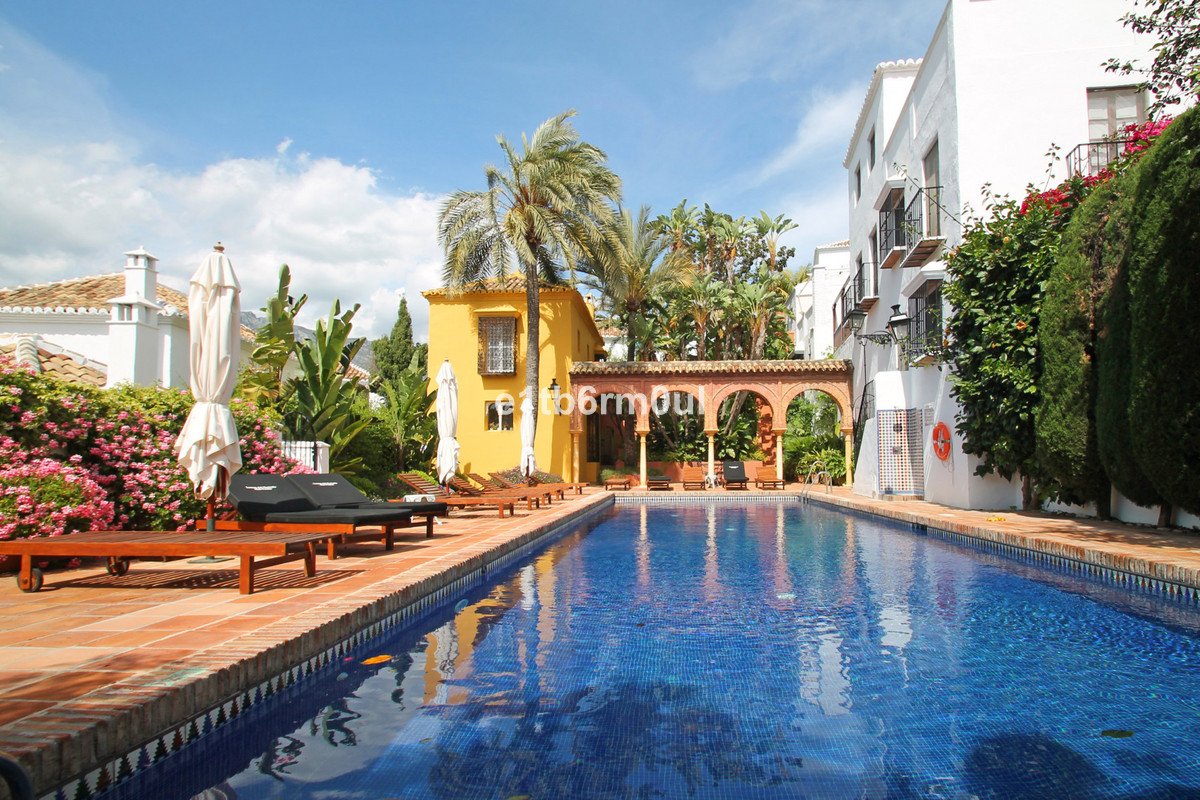 Outdoor swimming pool with lounge chairs, umbrellas, palm trees, and surrounding buildings on a sunny day.