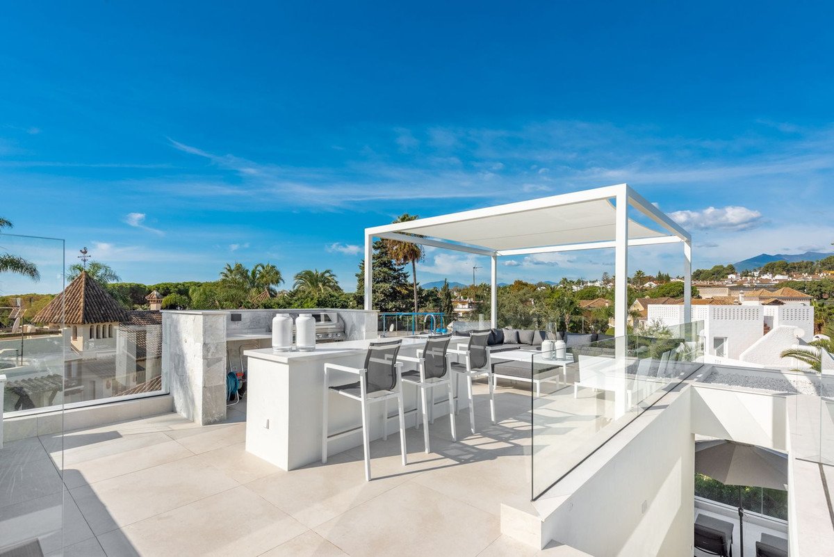 Modern rooftop terrace with an outdoor kitchen, bar, lounge area, and scenic views of hills and blue sky.